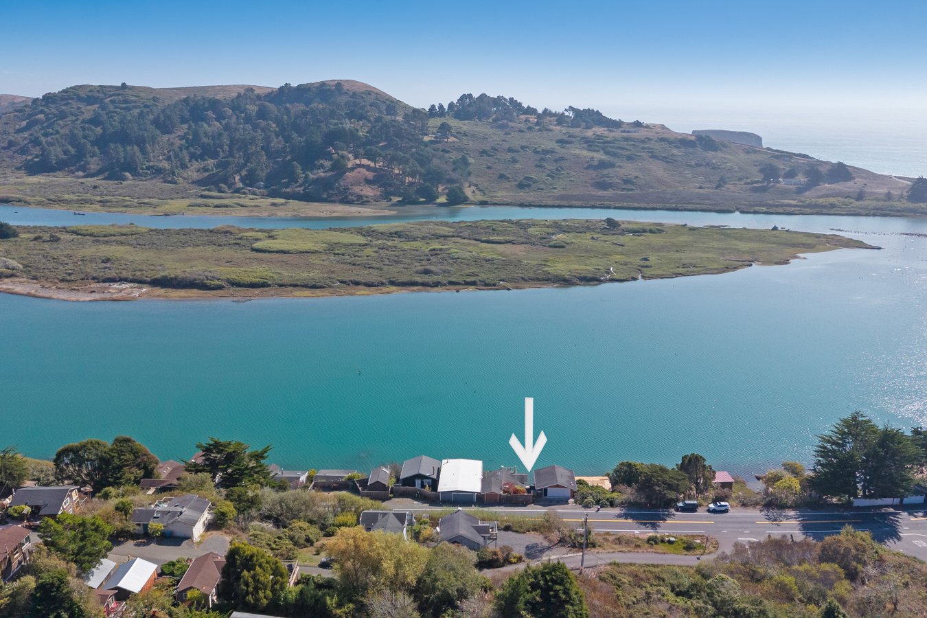 Aerial view of a lake with houses along the shore, with green hills and hills in the background under a clear blue sky.