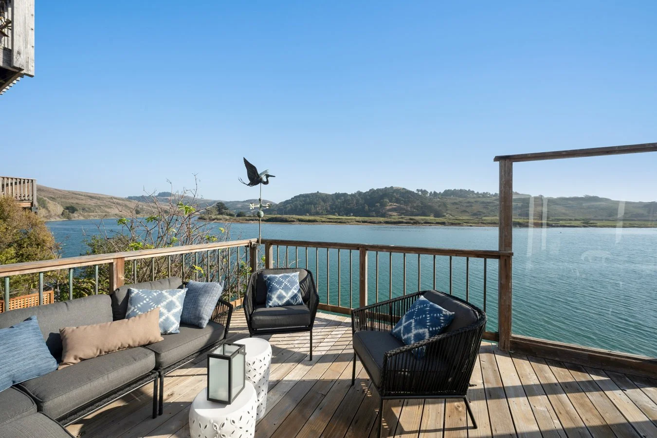 A wooden deck overlooking a body of water with hills in the distance. The deck has outdoor seating with cushions, a small white side table, and a weather vane shaped like a heron.