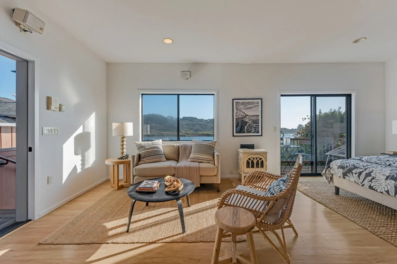 Living room with a beige sofa, black coffee table, woven chairs, and a sliding glass door leading to a balcony with outdoor seating. Windows show a hillside and water view, with minimalistic decor and natural lighting.