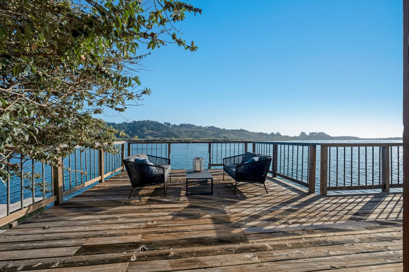 A wooden deck overlooking a body of water with two black chairs and a small table between them, surrounded by a railing, with a tree on the left and hills in the background, under a clear blue sky.