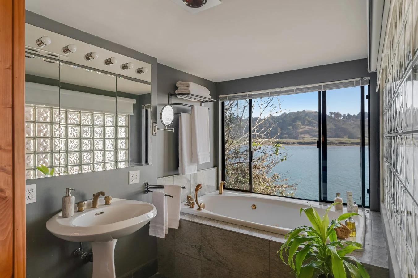 Bathroom with large window overlooking a lake and hills, featuring a bathtub, a sink with gold fixtures, a mirror, towels, and a potted plant.