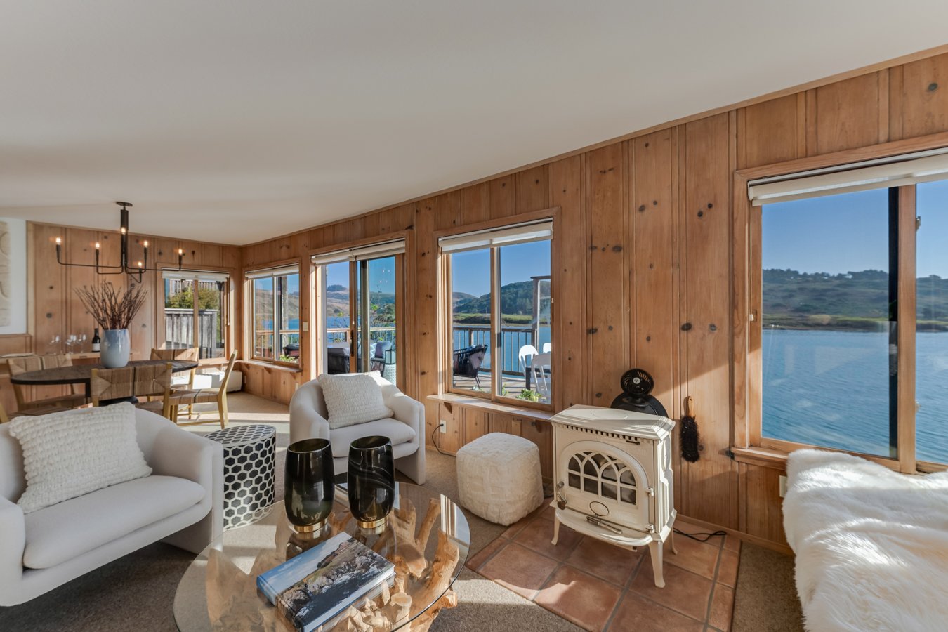 Living room with wooden panel walls, large windows overlooking a lake, white sofas, a coffee table, and decor.