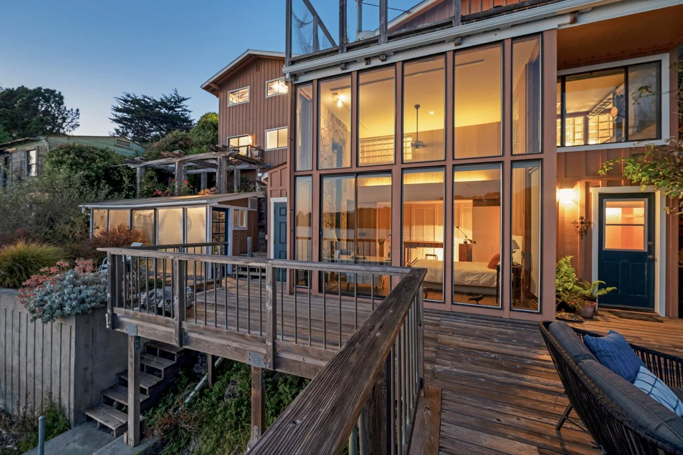 A multi-story house with large glass windows and a wooden deck illuminated by interior lights at dusk, surrounded by greenery.