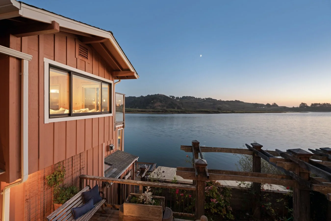 A lakeside house with large windows, a small dock, and a garden, during dusk or dawn with the moon visible in the sky.