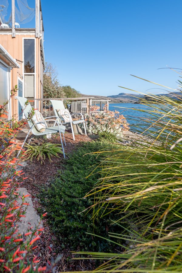 Balcony overlooking the water with patio chairs and plants, near a building with large windows.