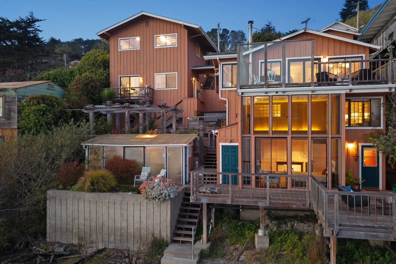 Multi-level wooden house with large glass windows and outdoor decks, surrounded by trees and plants at dusk.