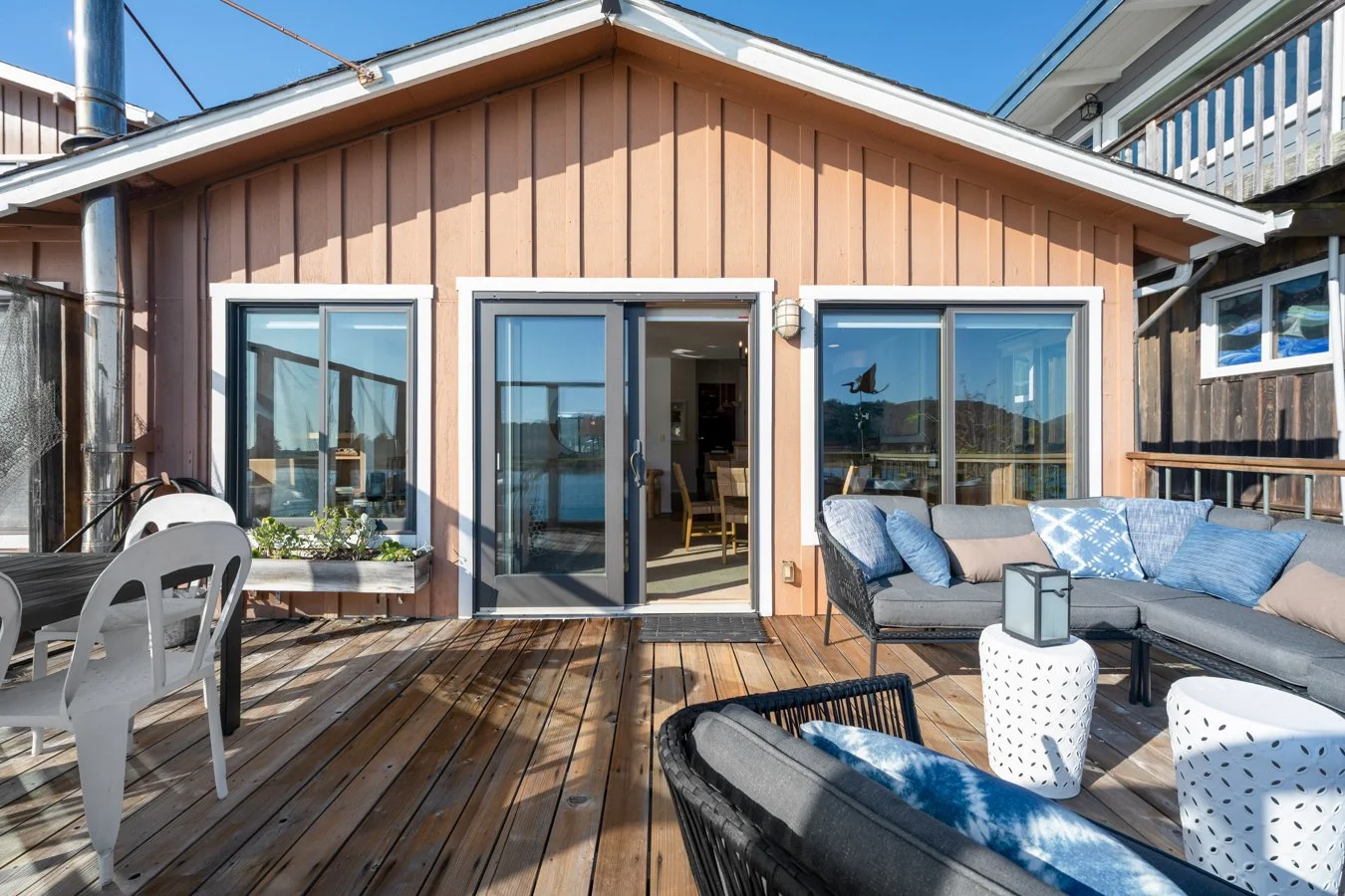 Outdoor wooden deck with a sectional sofa and seating area, view of a house with sliding glass doors and windows, blue sky, and mountain landscape in the background.