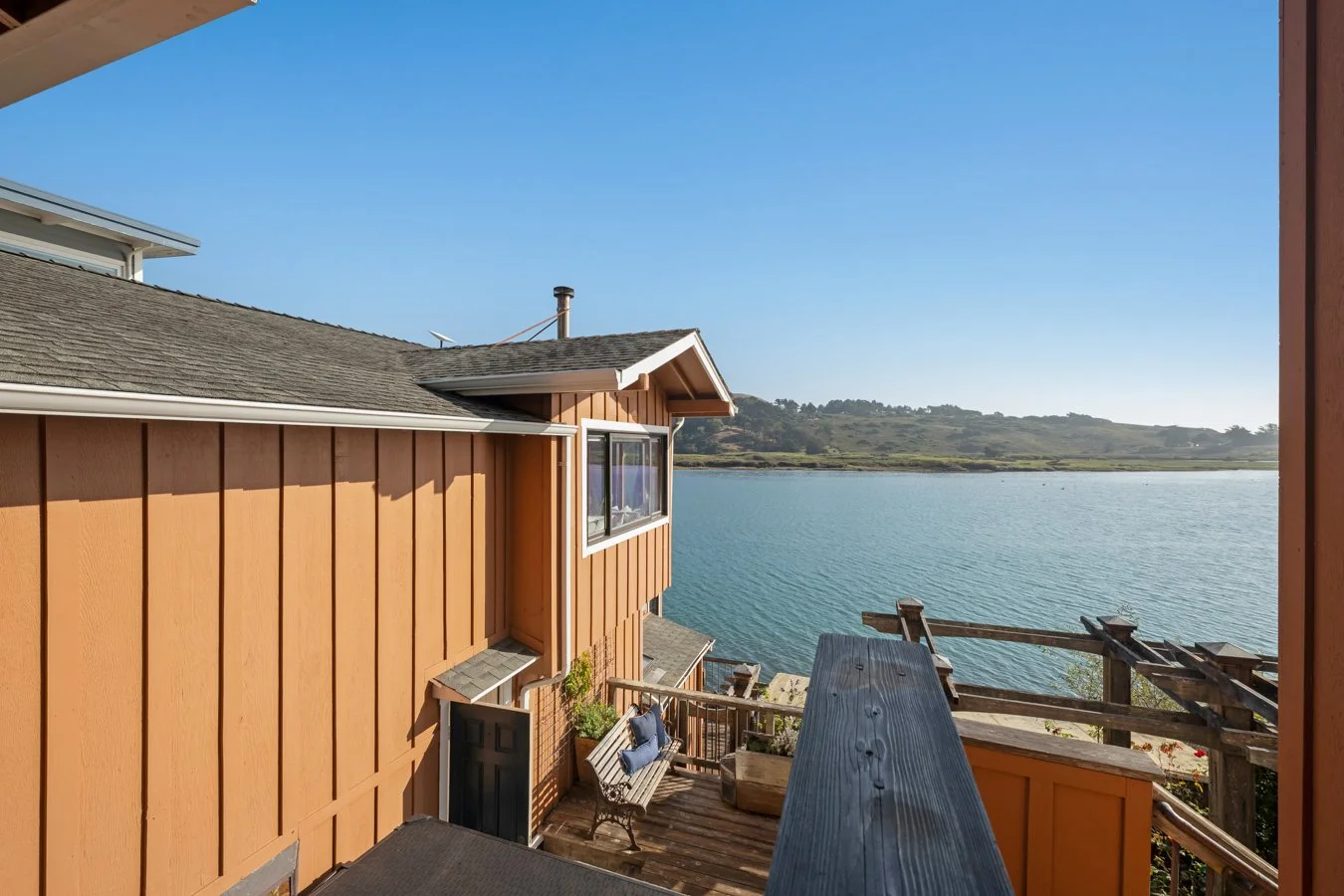 View from a balcony overlooking a large body of water with a distant shoreline and hilly landscape under a clear blue sky. The balcony has wooden railings, a bench with pillows, and part of a house with orange siding.