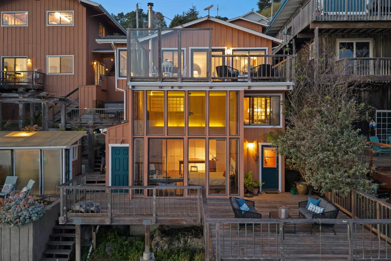 A multi-story house with large windows and multiple outdoor decks in a residential neighborhood during dusk. The house has warm interior lighting visible through the windows, with some furniture and decor inside. Surrounding neighboring houses are al