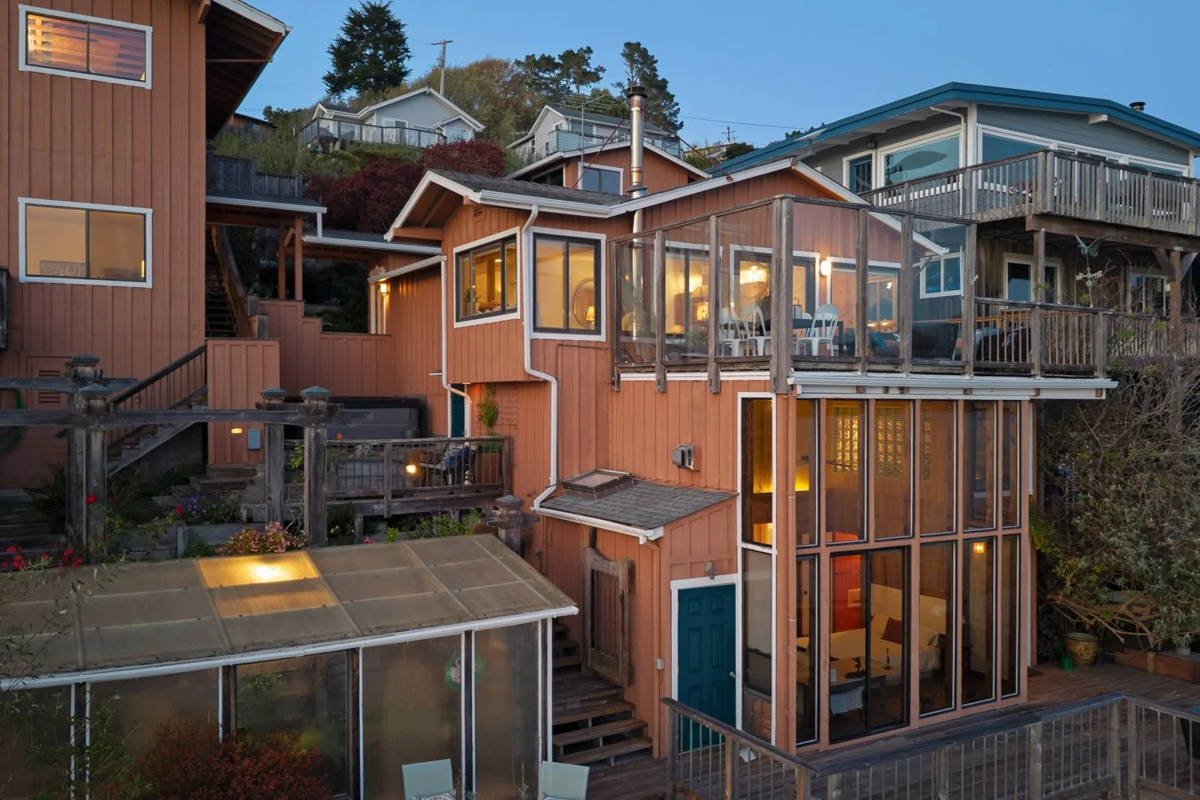 Multi-level wooden house with large glass windows and a balcony, situated on a hillside, with neighboring houses in the background during dusk.
