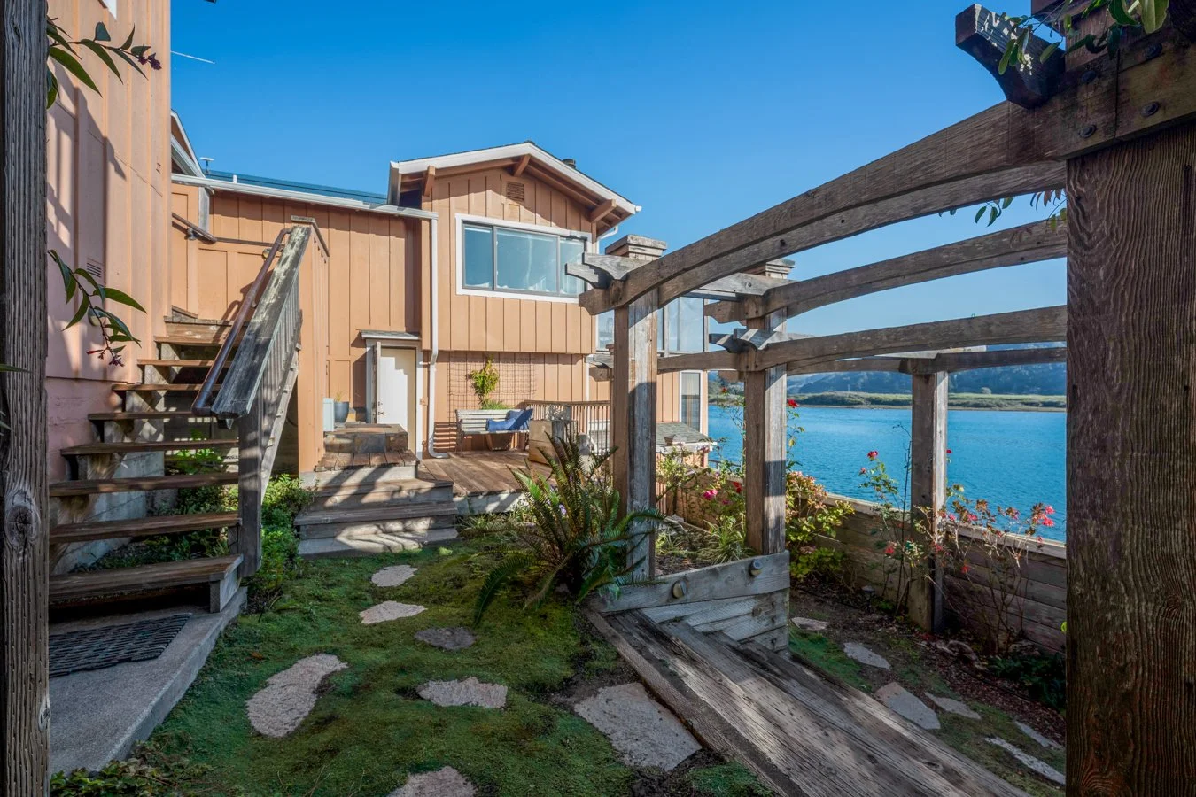 View of a house with a wooden deck near a body of water, featuring stairs, a garden with flowers, and a clear blue sky.
