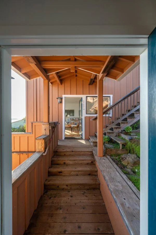 View of a small balcony or porch area with wooden flooring and orange siding, leading to an interior room with a couch, and an outdoor staircase with greenery on one side.