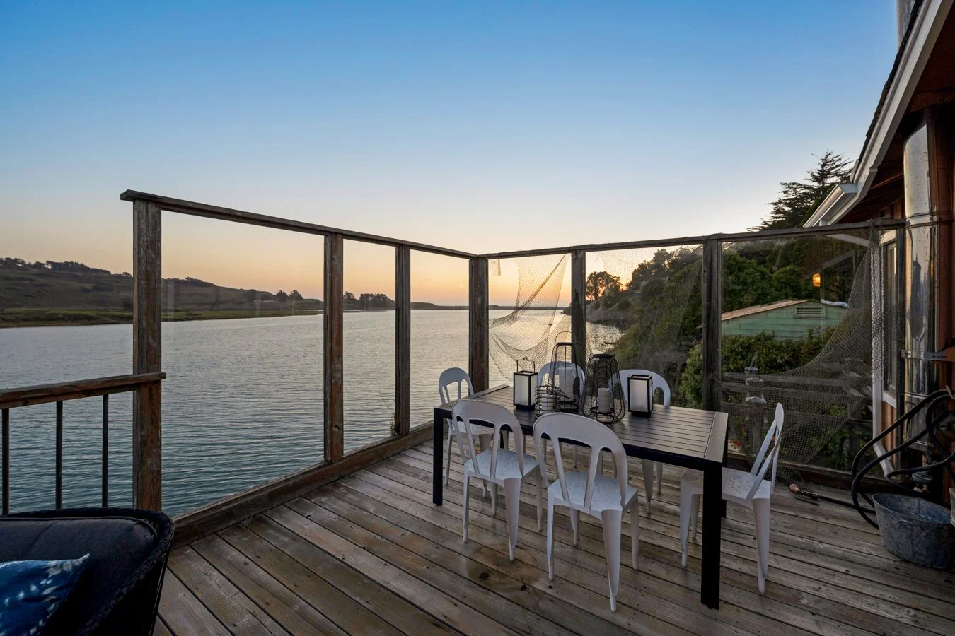 A wooden balcony overlooking a body of water at sunset, with a black dining table, white chairs, and lanterns, surrounded by a railing with a mesh screen.