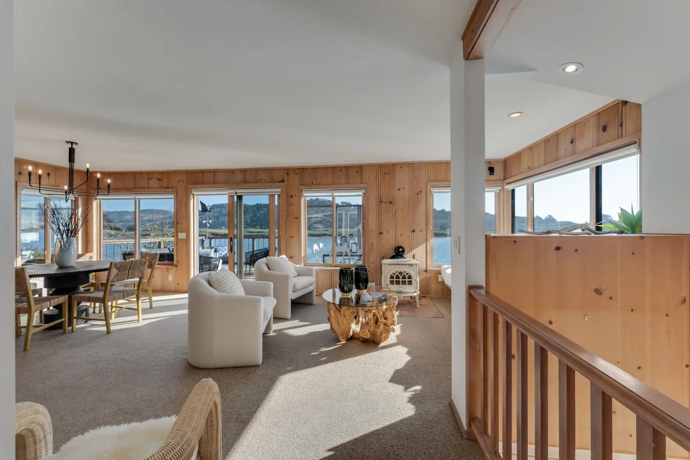 Living room with large windows overlooking a lake, white armchairs, a wooden coffee table, and a dining area with a chandelier.