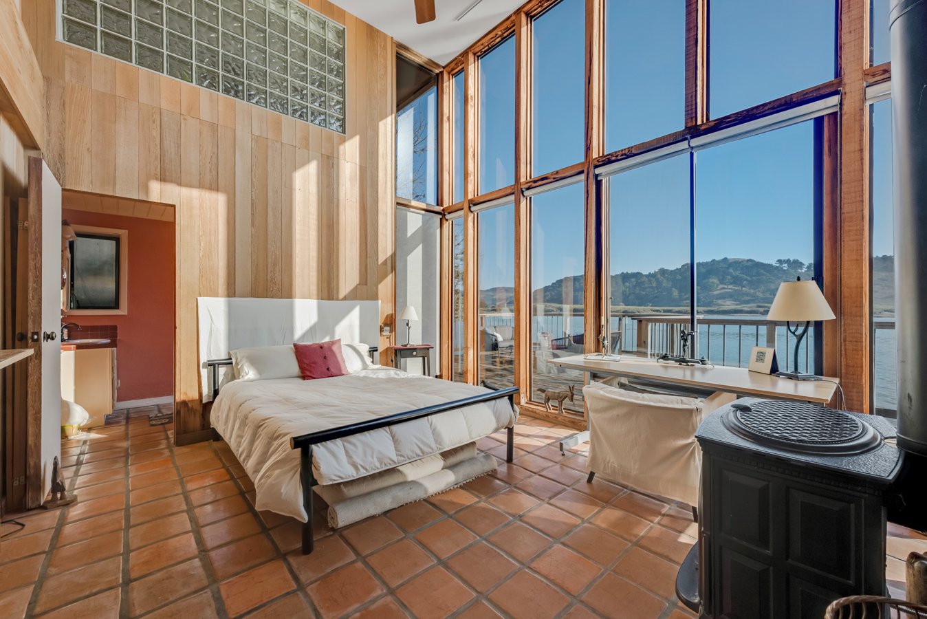 A bedroom with large floor-to-ceiling windows overlooking a body of water and hills, featuring terracotta tile flooring, a bed with white bedding and a red pillow, a white desk with a lamp and picture frame, and a black stove in the corner.