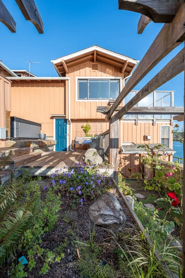 A two-story house with peach-colored siding and a blue front door, surrounded by a garden with flowers and rocks, and a blue sky overhead.