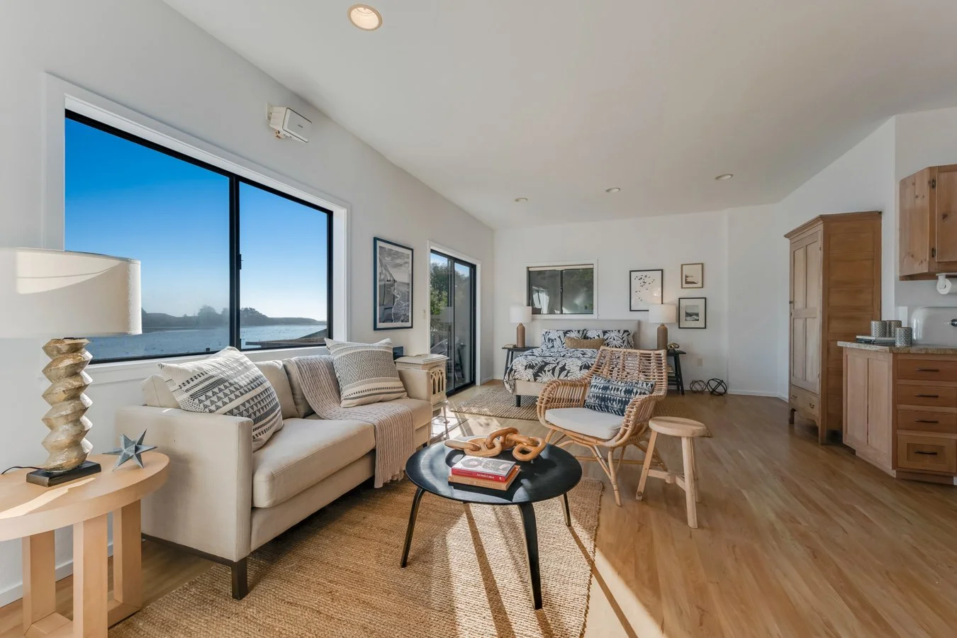 Bright living room with large windows overlooking water, white sofa with patterned pillows, black coffee table with books and decor, wicker chairs, and coastal decor, hardwood floors
