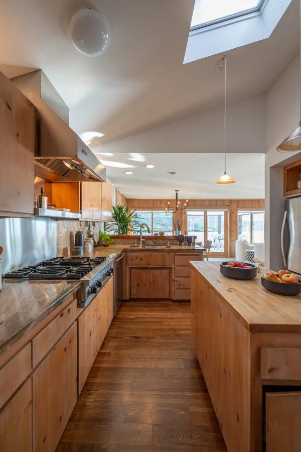 Cozy kitchen with wood cabinets, countertop appliances, and large windows revealing a bright living area with dining table and chairs.