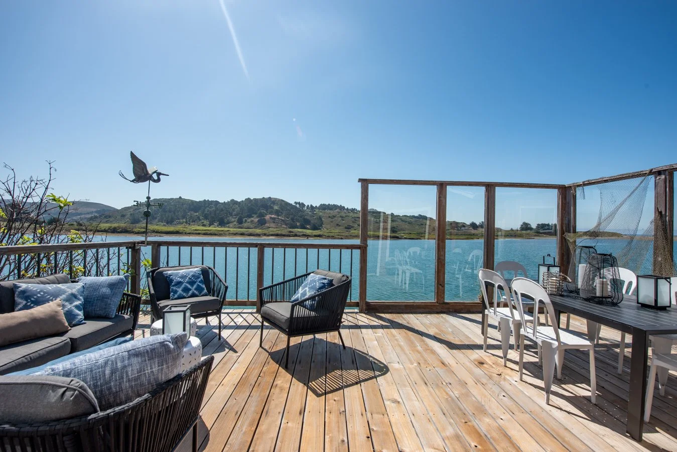 Wooden deck overlooking a lake with outdoor furniture, including a sofa, chairs, and a dining table, under a clear blue sky.
