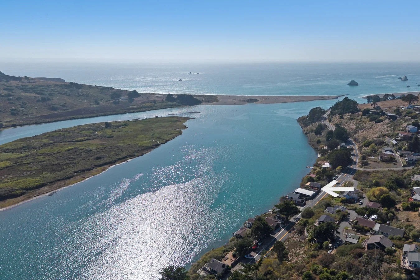 Aerial view of a coastal area with a river flowing into the ocean, houses on hills, green vegetation, and a clear blue sky.