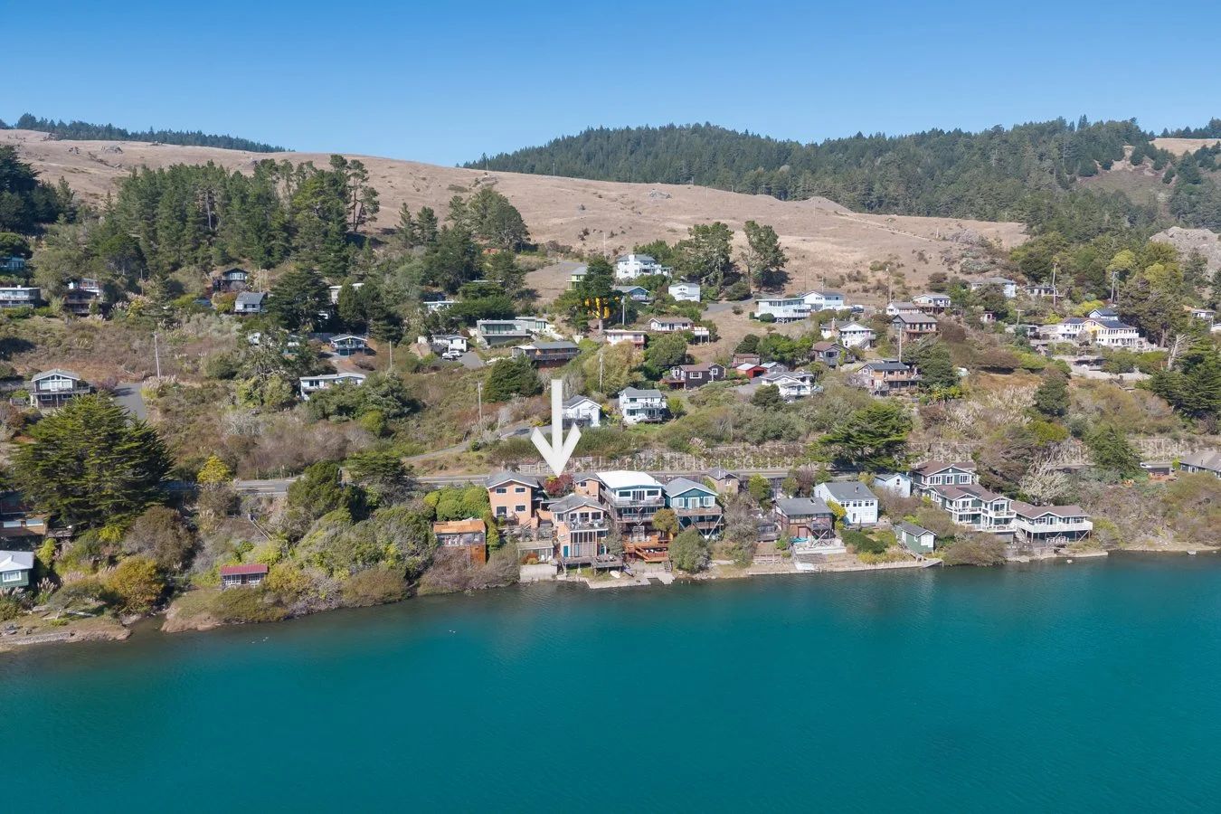 Aerial view of a lakeside neighborhood with houses on a hillside and a body of water in the foreground. An arrow points to a specific house.