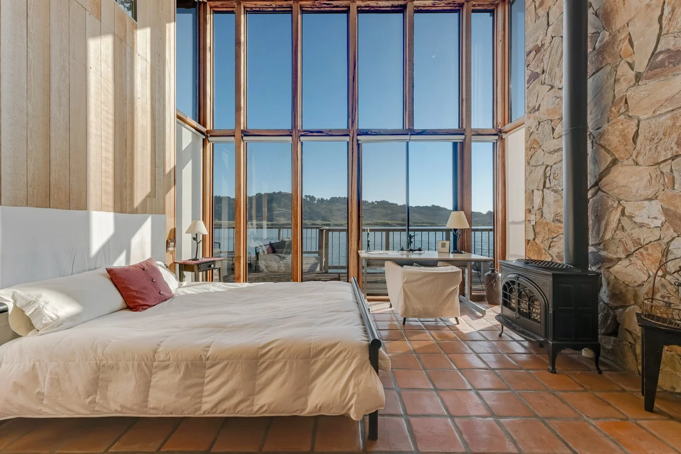 Bedroom with large floor-to-ceiling windows overlooking a body of water and distant hills, a bed with white sheets and a pink pillow, a small desk with a lamp, a chimney, and terracotta tile flooring.