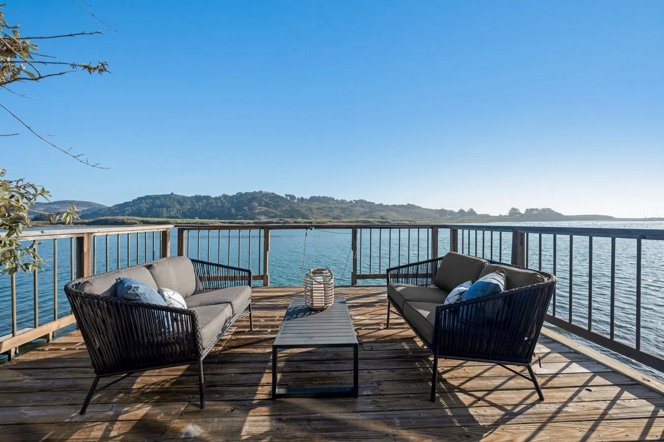 Outdoor wooden deck overlooking a body of water with two black framed sofas and a black coffee table, under clear blue sky.