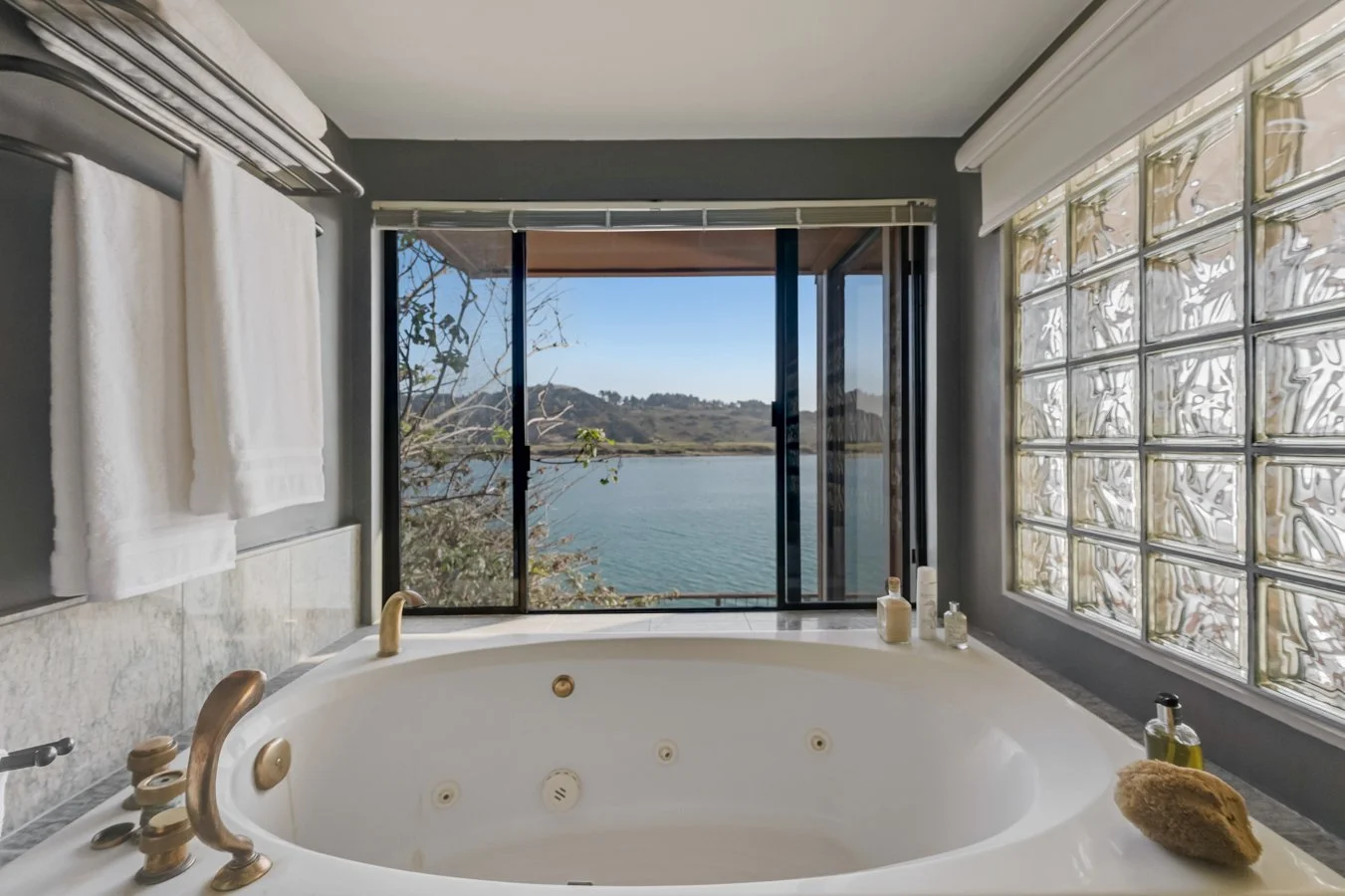 Bathroom with large window overlooking a lake and hills, featuring a white bathtub with gold fixtures, towels, and toiletries.