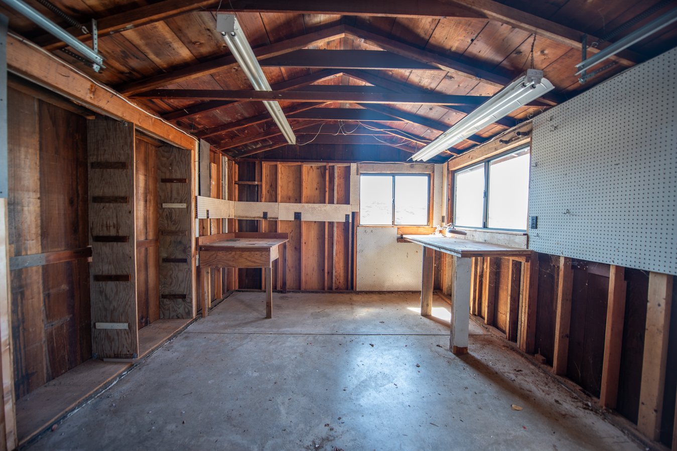 Wooden shed interior with unfinished walls, concrete floor, two workbenches, and windows.