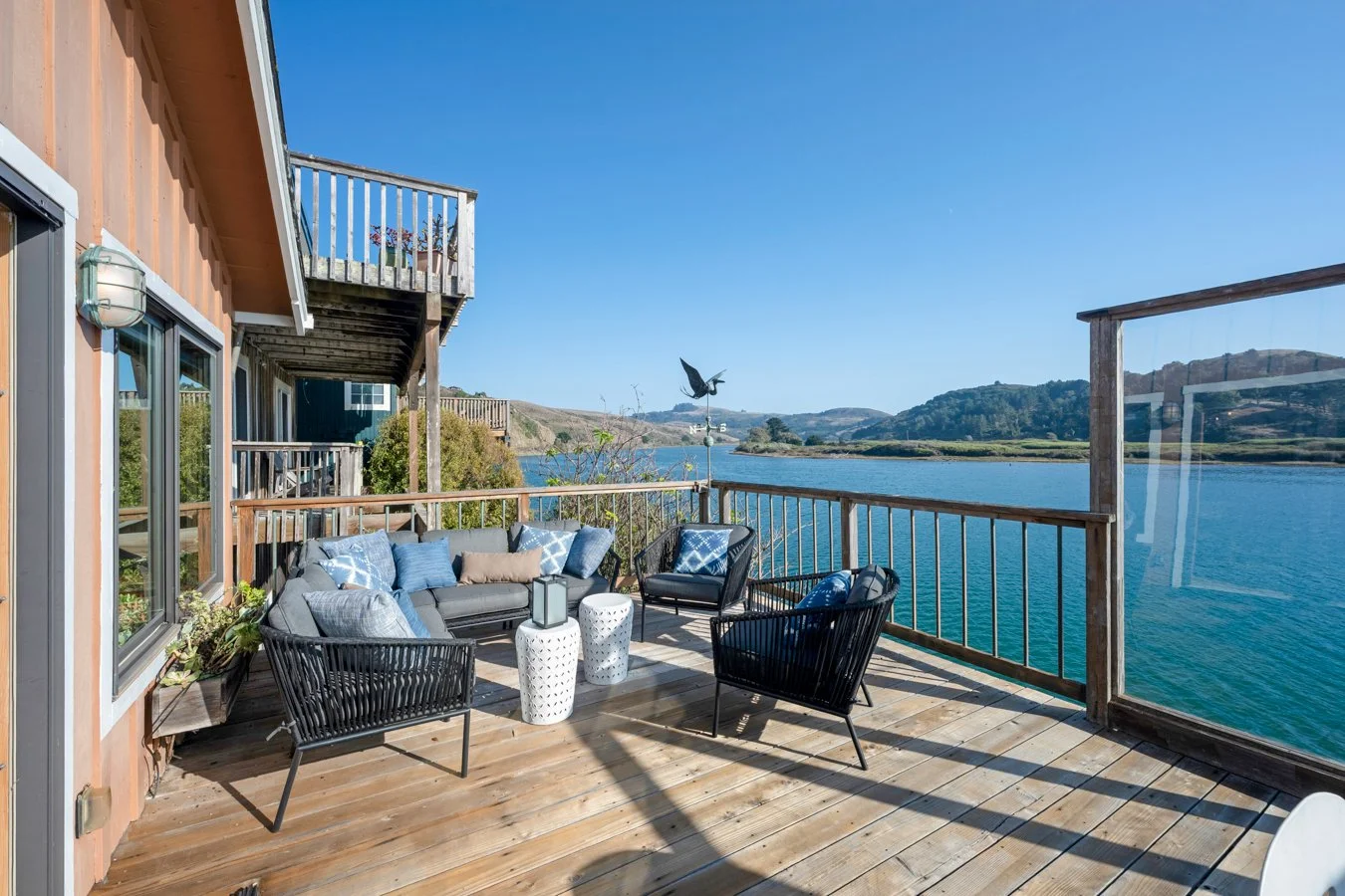 A wooden deck overlooking a body of water with hills in the background. The deck has a gray sofa with blue and beige pillows, black chairs, two white side tables, and a decorative metal bird with wings spread.