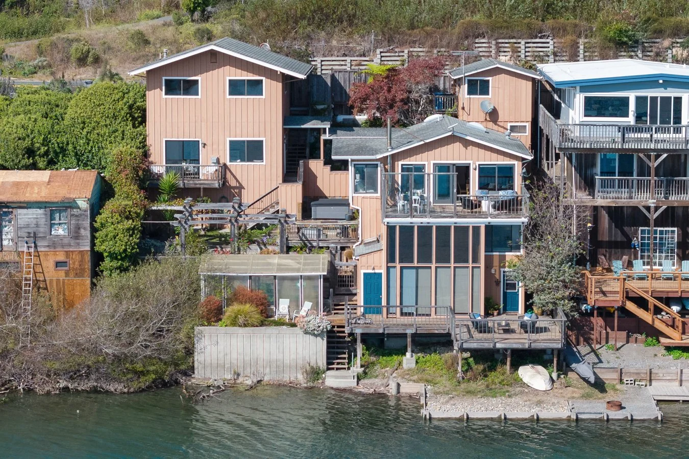 A row of waterfront houses on a hillside, featuring multiple decks, large windows, and outdoor furniture, with the water in the foreground.