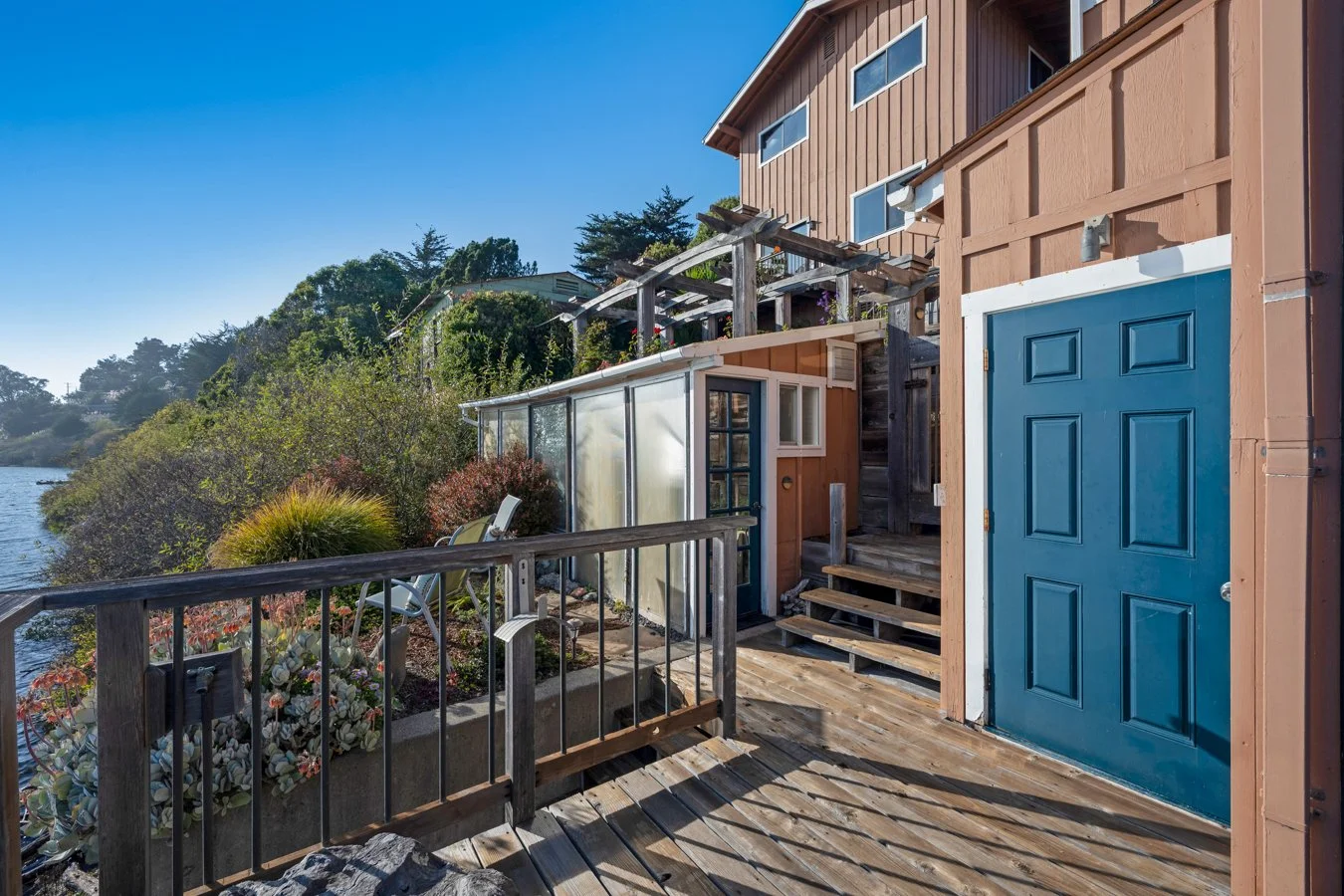 A wooden deck of a house beside a body of water with lush greenery in the background. The deck has a blue door, stairs, and a small greenhouse or porch area with a window.