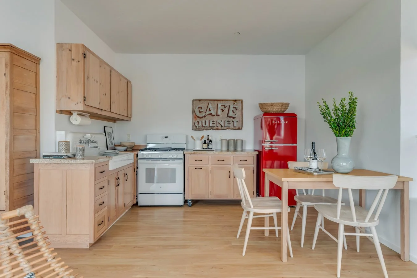 A kitchen with light wood cabinets, white stove, red vintage refrigerator, and a small dining table with white chairs. Decor includes a sign reading 'Cafe Queret,' a large plant in a white vase, and various kitchenware.