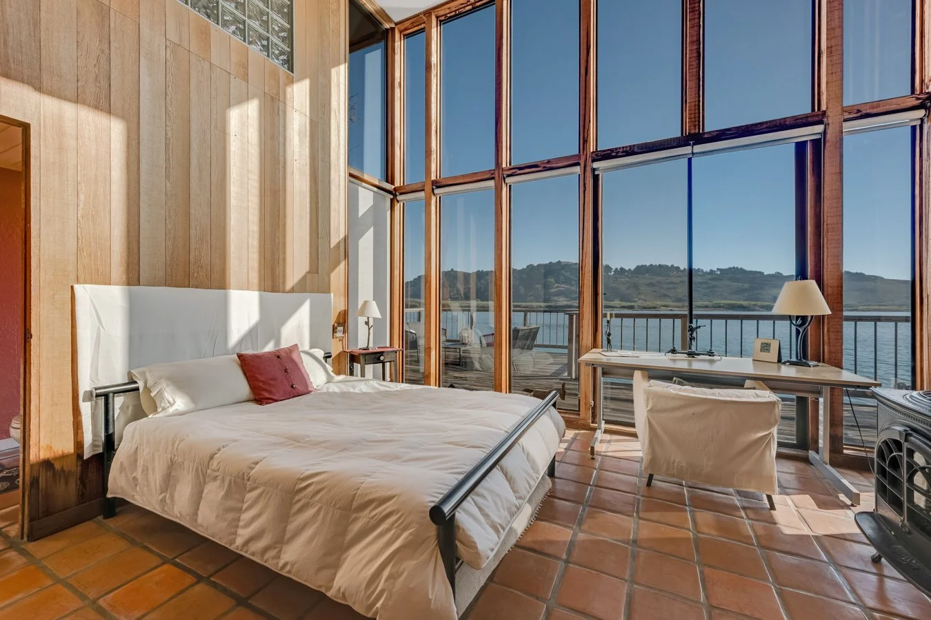 Bedroom with large glass windows overlooking a body of water and hills, with a bed, side tables, lamps, and a desk.