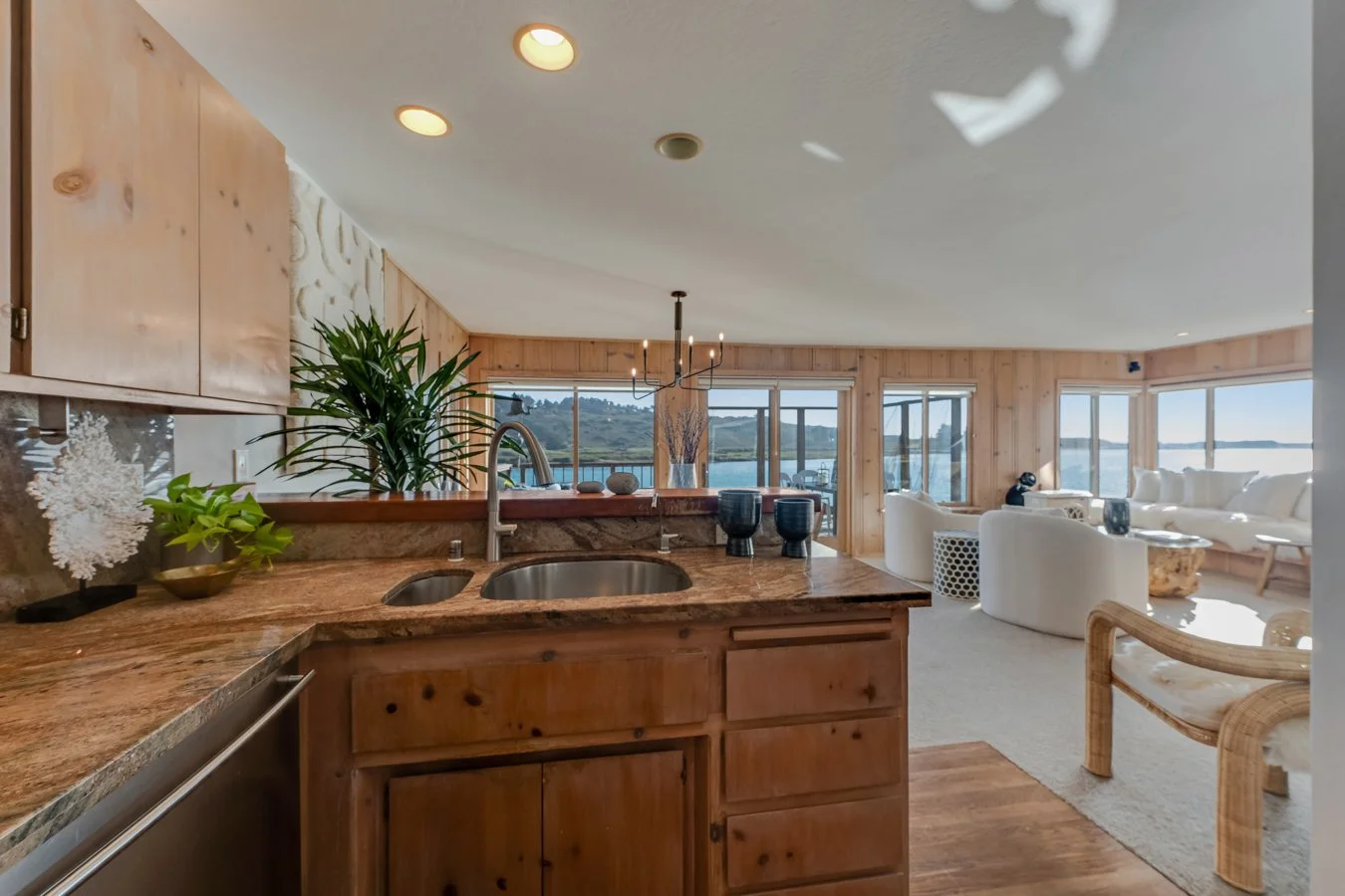 Open-concept living room and kitchen with ocean view, wood-paneled walls, white furniture, and large windows.