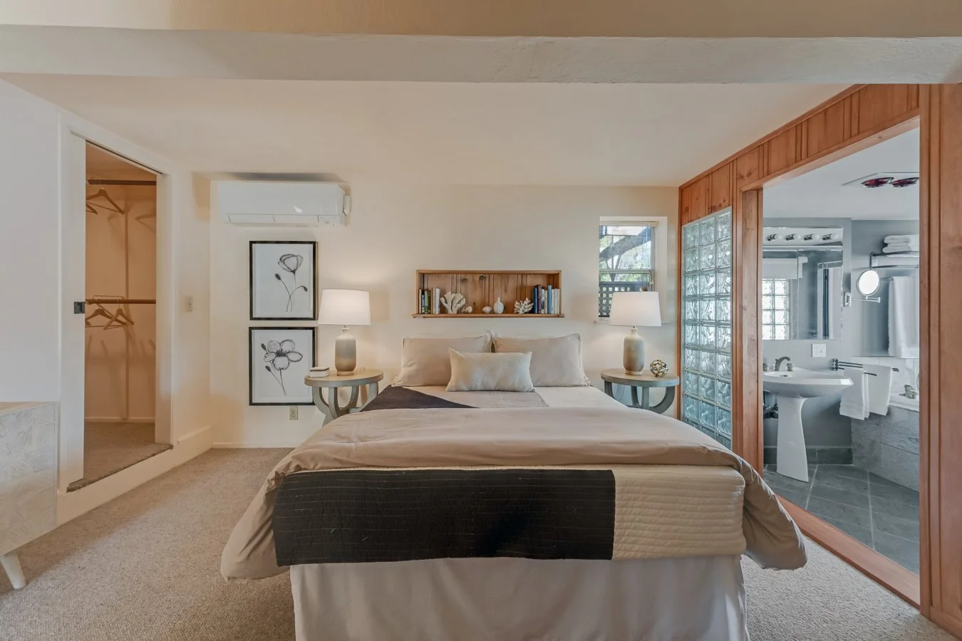 A cozy hotel bedroom with a bed flanked by two nightstands with lamps, a wall shelf above the bed, and a partial view of a bathroom through a glass block wall.