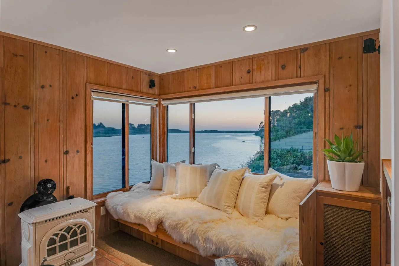A cozy seating nook with a plush, fluffy cushion and multiple pillows, surrounded by wood-paneled walls, large windows showcasing a river view, a small white heater, a potted plant, and soft indoor lighting.