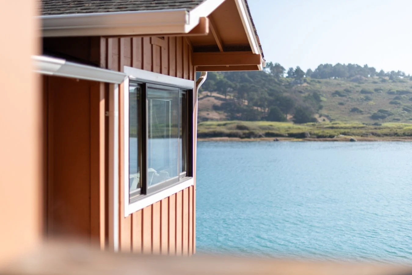 Side view of a red wooden house with a large window, overlooking a body of water with hills and trees in the background.
