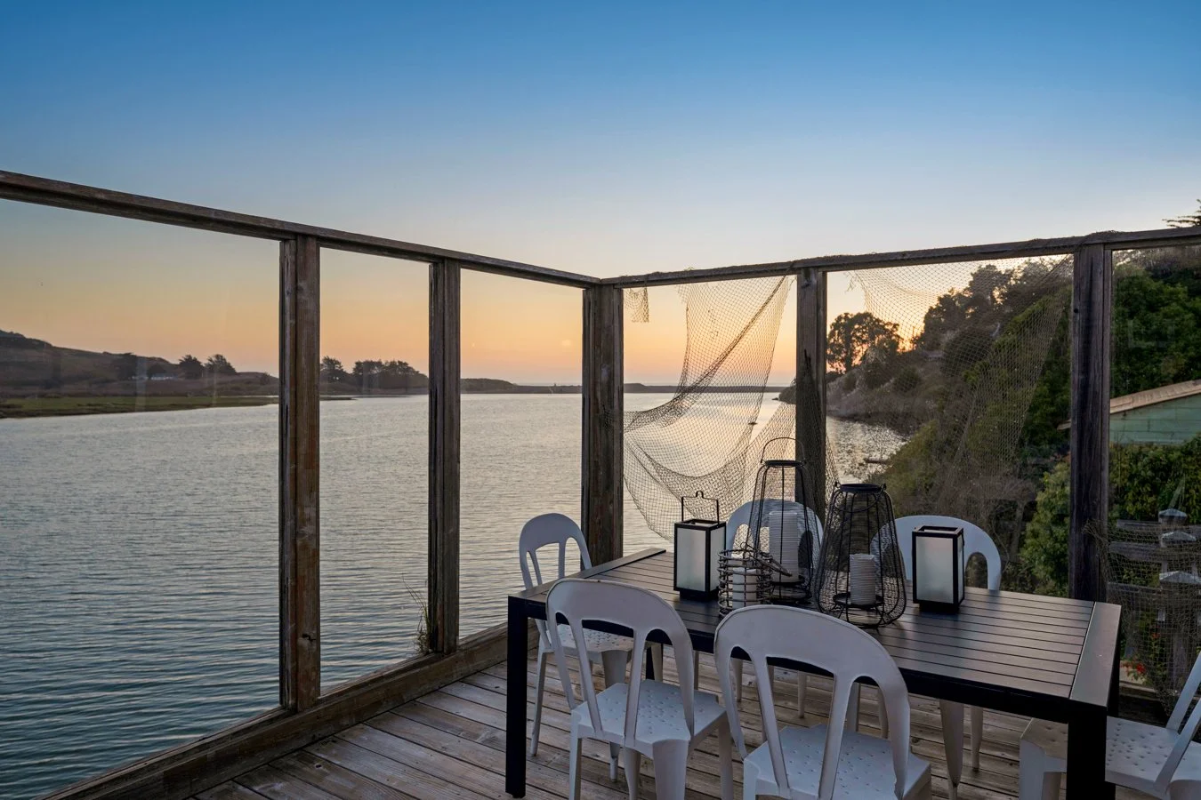 Outdoor dining area on a wooden deck overlooking a river at sunset, with lanterns and chairs, surrounded by a wooden railing and some fishing nets hanging on the fence.