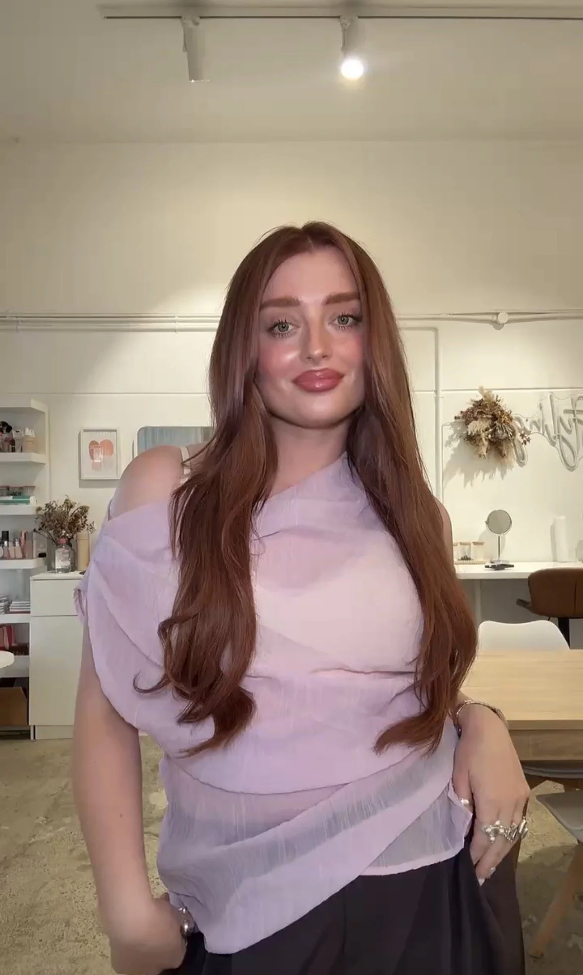 A woman with long red hair and light makeup wearing a pink off-shoulder top and black pants, standing in a room with white walls, shelves with decor, and a table in the background.