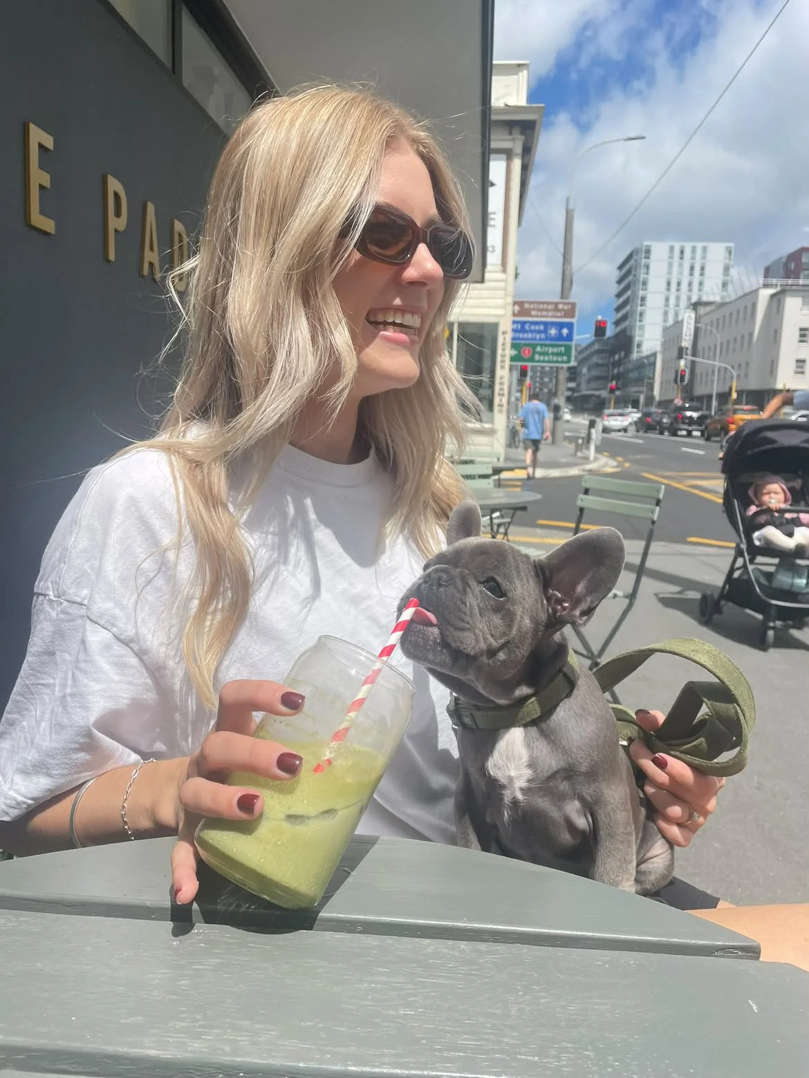 A woman with blonde hair, sunglasses, and a white shirt is sitting outdoors at a table, holding a green smoothie with a red and white striped straw. A French bulldog puppy is licking the straw while being held by the woman, and a baby is in a strolle
