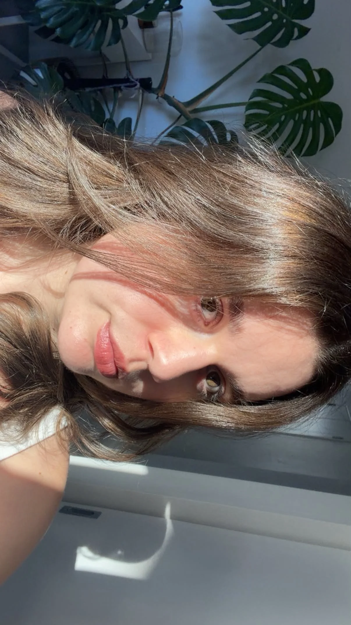 A woman with light brown hair lying on her side, with a plant with large green leaves in the background.