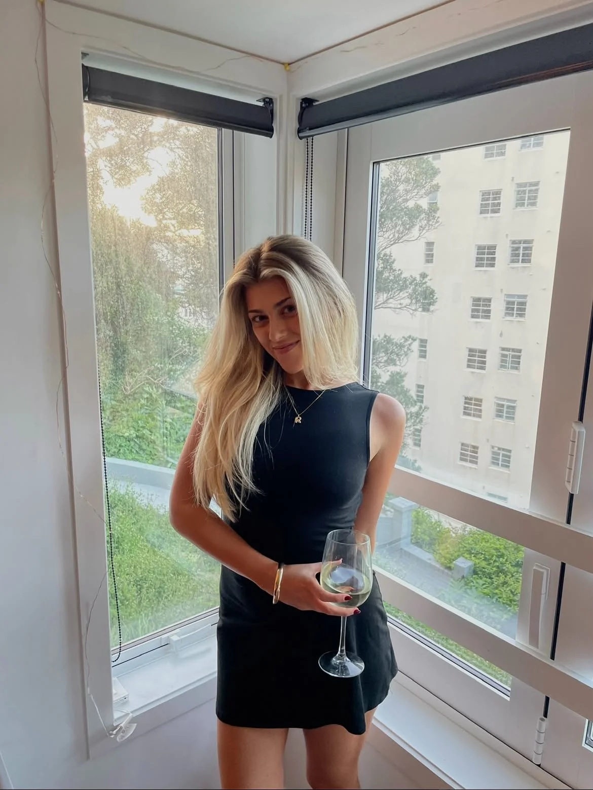 A woman with long blonde hair wearing a black sleeveless dress and holding a glass of white wine, standing in front of large windows with a view of a cityscape and greenery outside.