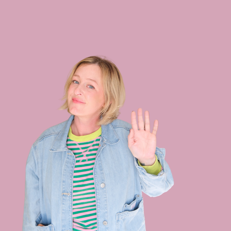 A woman with blonde hair, wearing a denim jacket over a colorful striped shirt, is raising her hand in a gesture of greeting or saying stop, against a pink background.