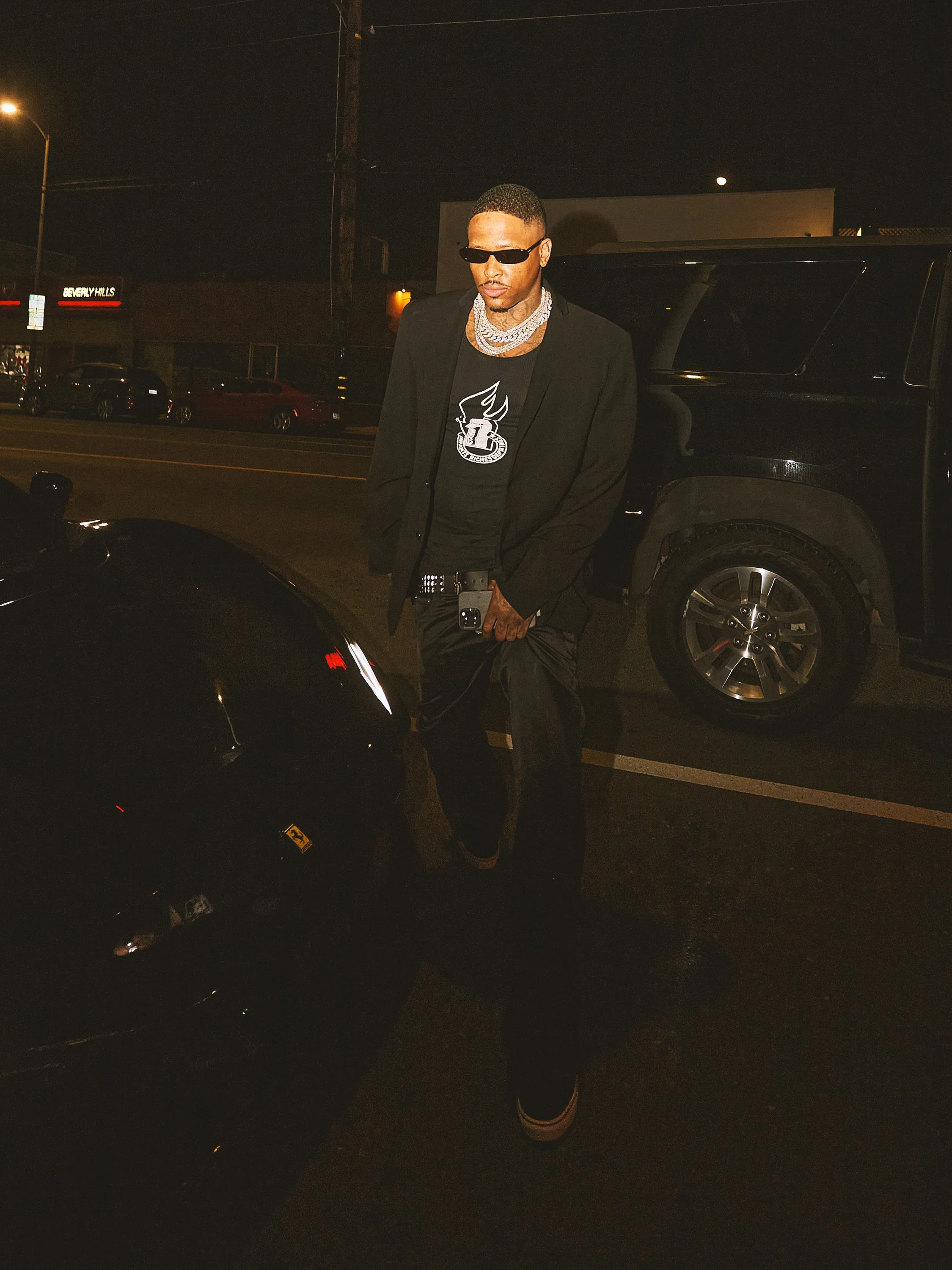 A young man wearing black sunglasses, a black blazer, layered necklaces, a black t-shirt with a white logo, and black pants, standing outdoors at night next to a black vehicle, with a neon sign reading 'Beverly Hills' in the background.