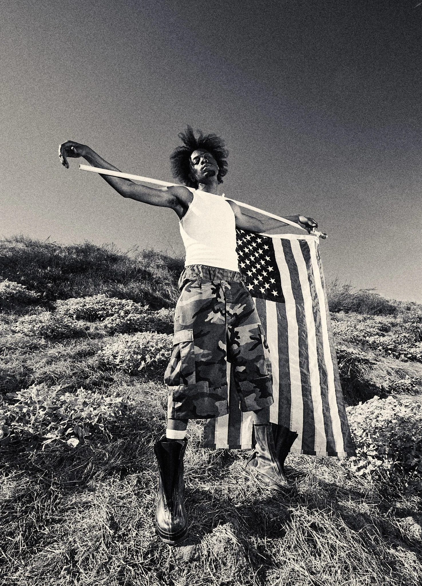 A person with curly hair stands in a field, holding an American flag over their shoulders and a stick in their mouth, wearing camouflage shorts and rain boots, in black and white photograph.