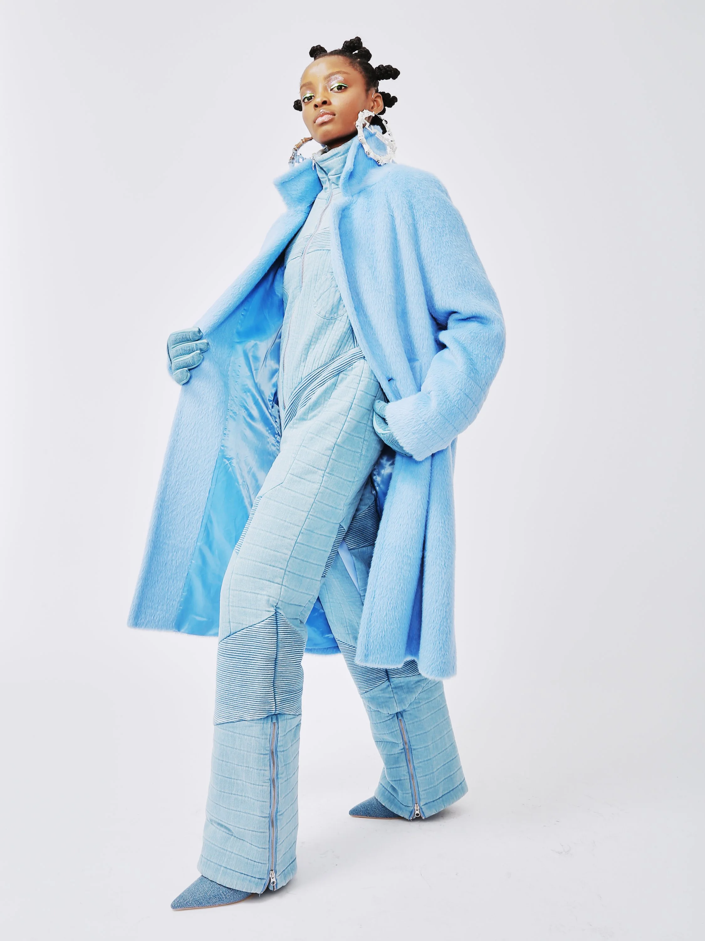 A woman wearing a light blue fur coat, matching light blue pants with zippers at the bottom, and pointed blue shoes. She accessorizes with large silver statement earrings and has makeup featuring bright green eyeshadow. She stands against a plain white background with a confident pose.