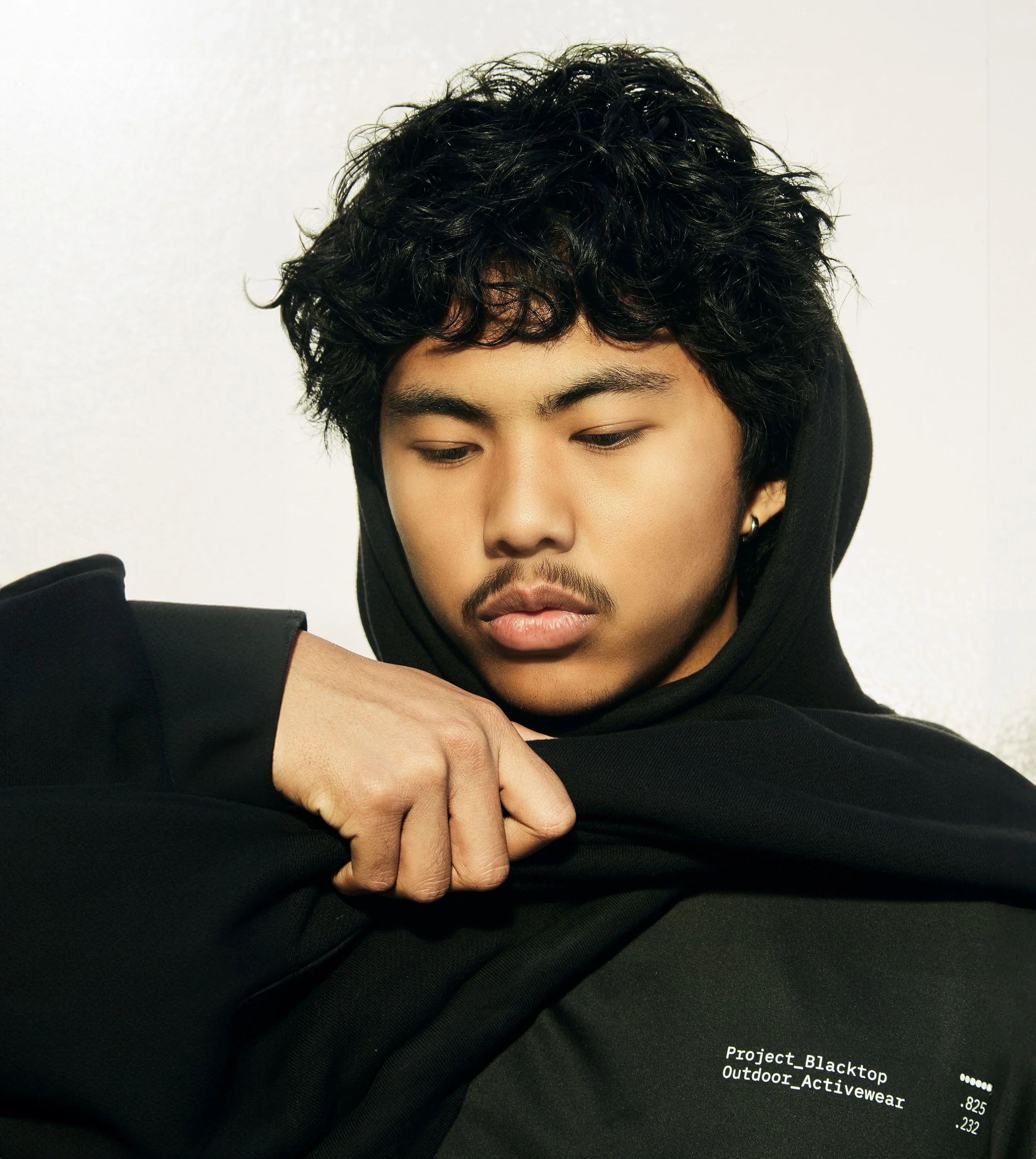 Young man with curly black hair wearing a black hoodie, looking down, against a white background, with partial text on the bottom right corner reading 'Project_Blacktop Outdoor_Activewear'.