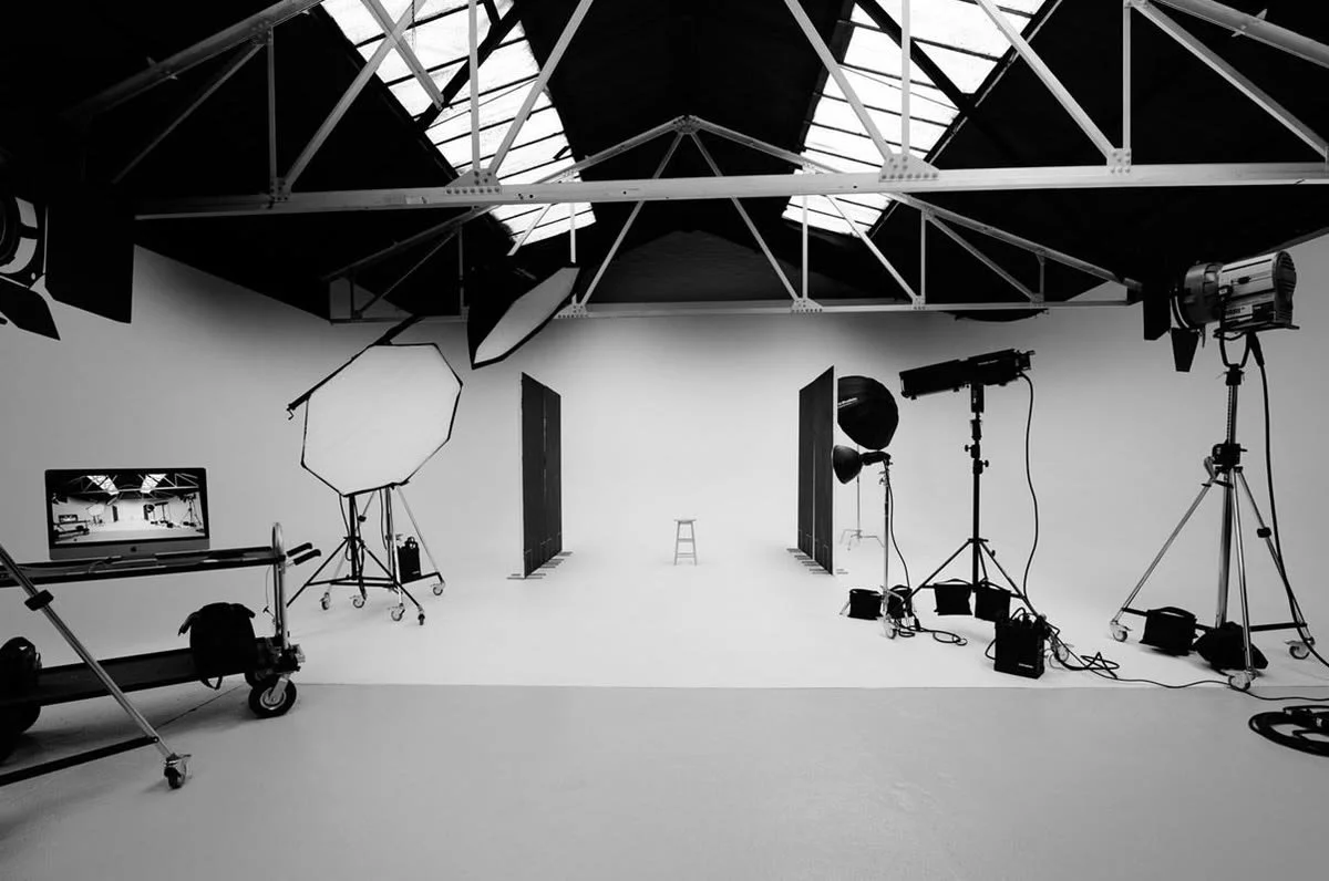 Photography studio with lighting equipment, a laptop on a cart, and a small stool in front of a plain white backdrop.