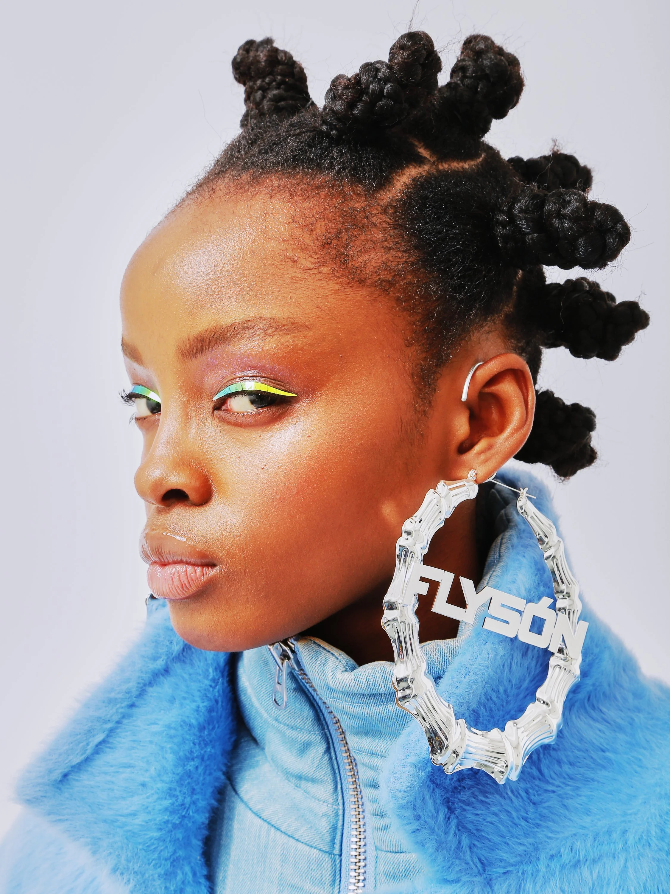A woman with dark skin and curly, twisted hairstyle wearing a bright blue, fluffy zip-up hoodie, silver earrings, and neon-colored eye makeup. She has small silver earrings and a large clear acrylic chain necklace with the word 'FUTURE' on it. The background is plain white.